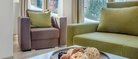 A modern living room with a sofa, armchair, and a table with a bowl of snacks. Large windows offer a view of the outdoors.