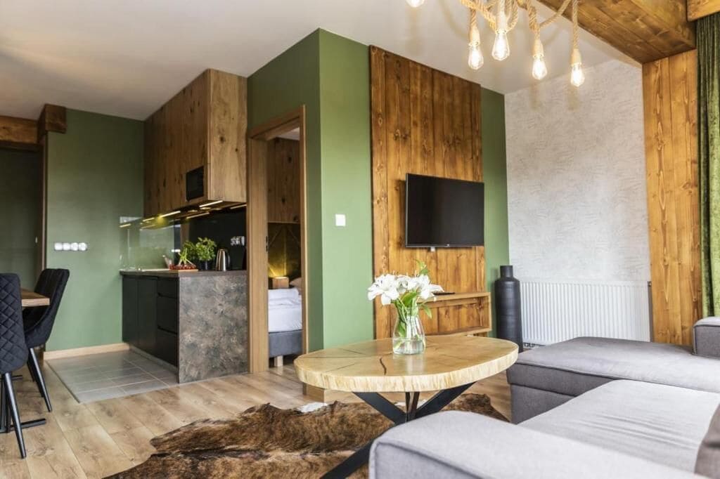 Ein modernes Wohnzimmer mit einer Couch, einem runden Holztisch, einem an einer Holzwand befestigten Fernseher und einer Küchenzeile mit Esstisch und Stühlen.