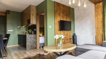 Ein modernes Wohnzimmer mit einer Couch, einem runden Holztisch, einem an einer Holzwand befestigten Fernseher und einer Küchenzeile mit Esstisch und Stühlen.