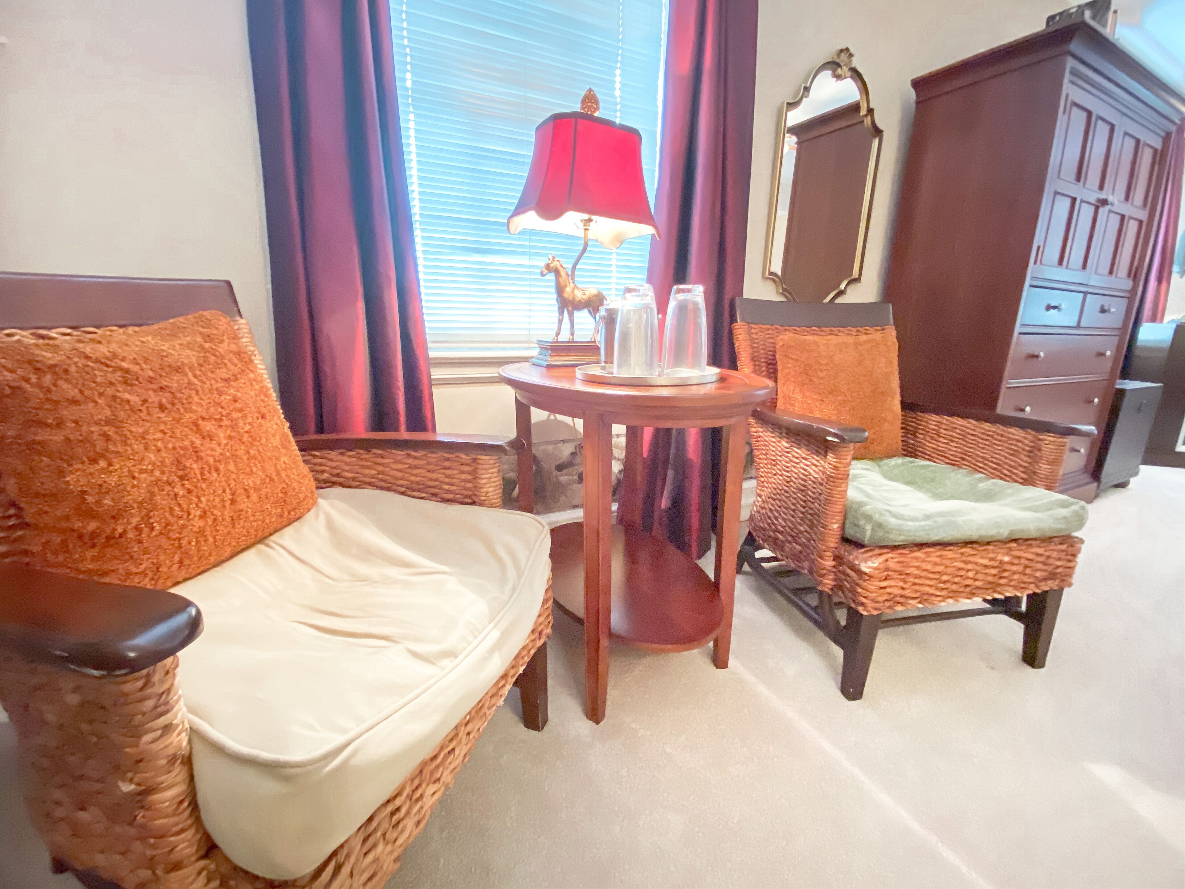 Een kamer met een rieten bank, een bijpassende fauteuil, een kleine ronde tafel met een rode lamp en een spiegel aan de muur.