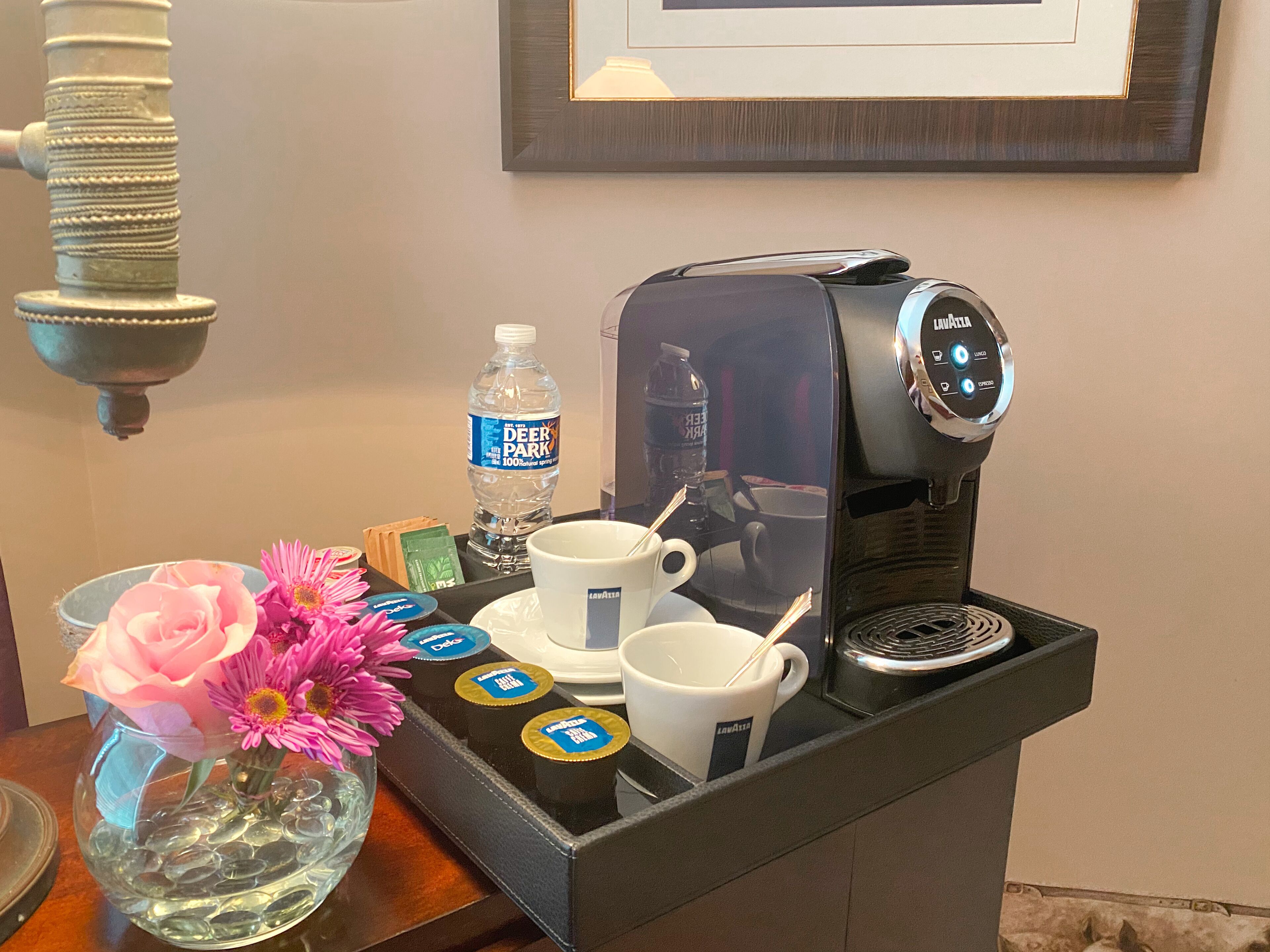 A coffee machine with a cup and saucer, a bottle of water, and a vase with flowers on a table.