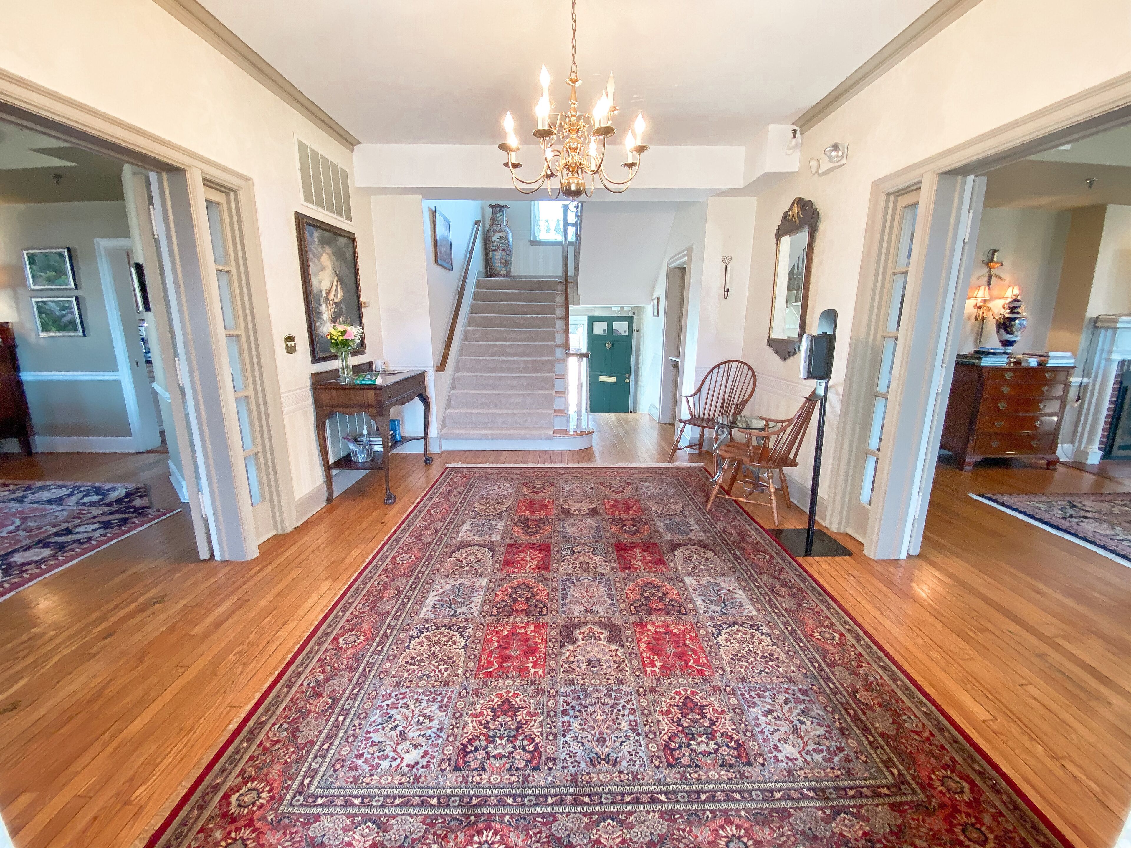 A spacious hallway with a large patterned rug, wooden flooring, a chandelier, and a staircase leading to another room.