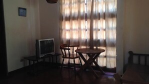 A room with a television on a stand, a round wooden table, two wooden chairs, and a window with sheer curtains. - Hotel Flor de Itabo (Sardinal)