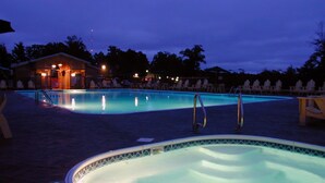 Zona de piscina con sillas de descanso y un jacuzzi, rodeada de árboles y un edificio iluminado.