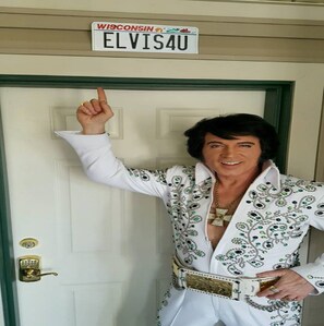 A person in a white suit with green and black embellishments standing in front of a door with a sign that reads "WISCONSIN ELVIS4U".