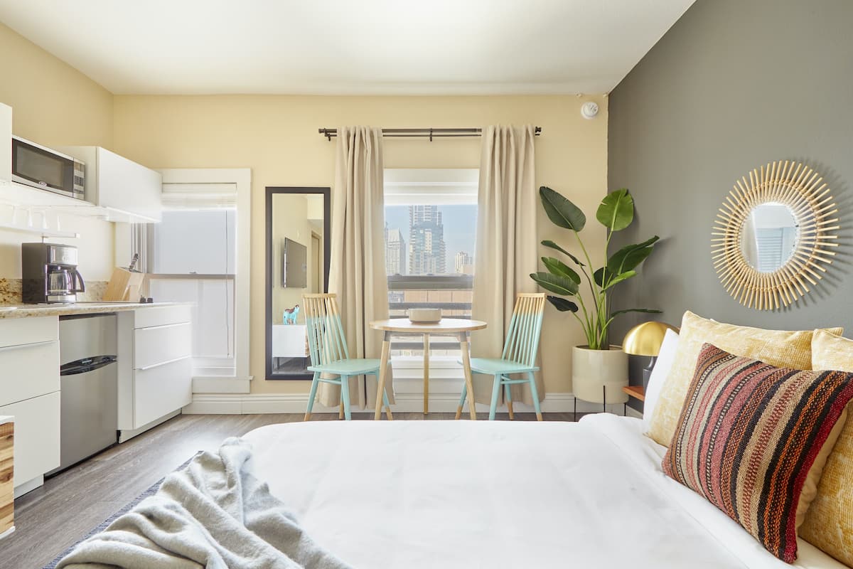 Ett hotellrum med en säng, ett matbord med stolar, ett köksutrymme och utsikt över stadsbilden.