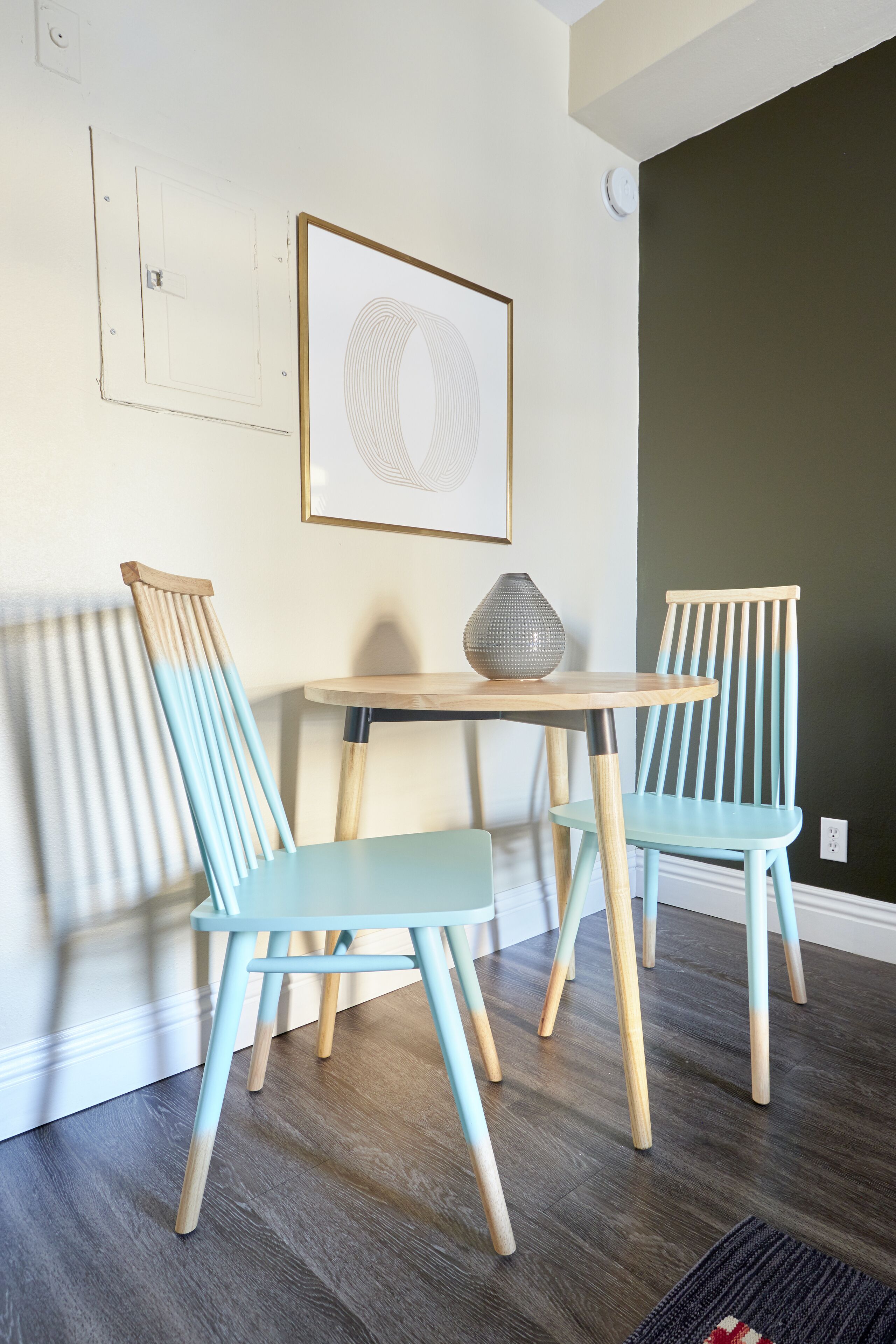 A room with two light blue chairs, a round wooden table, and a framed abstract artwork on the wall.