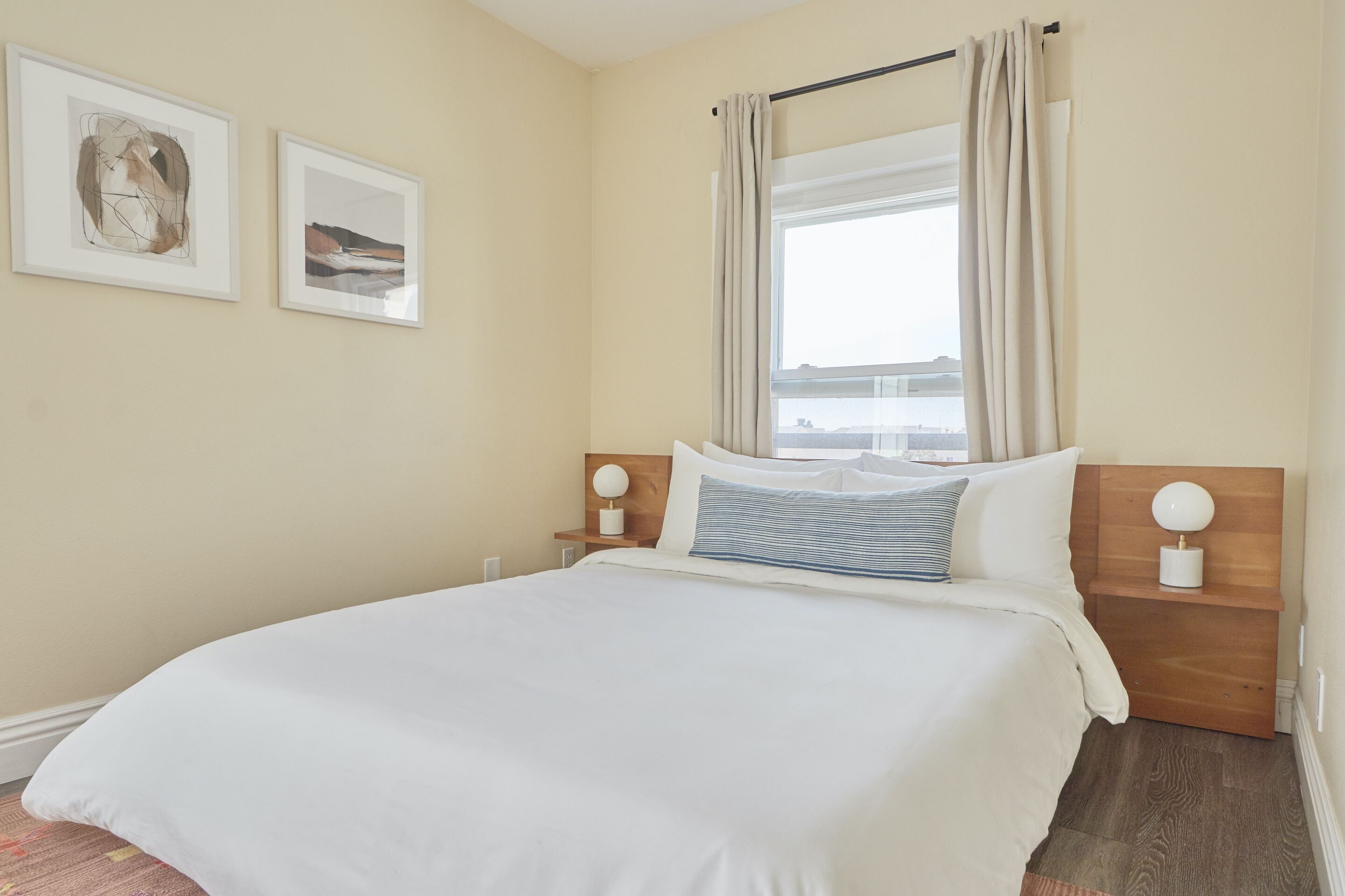 A neatly made bed with white linens, a wooden headboard, and two bedside tables with lamps. There are two framed pictures on the wall.