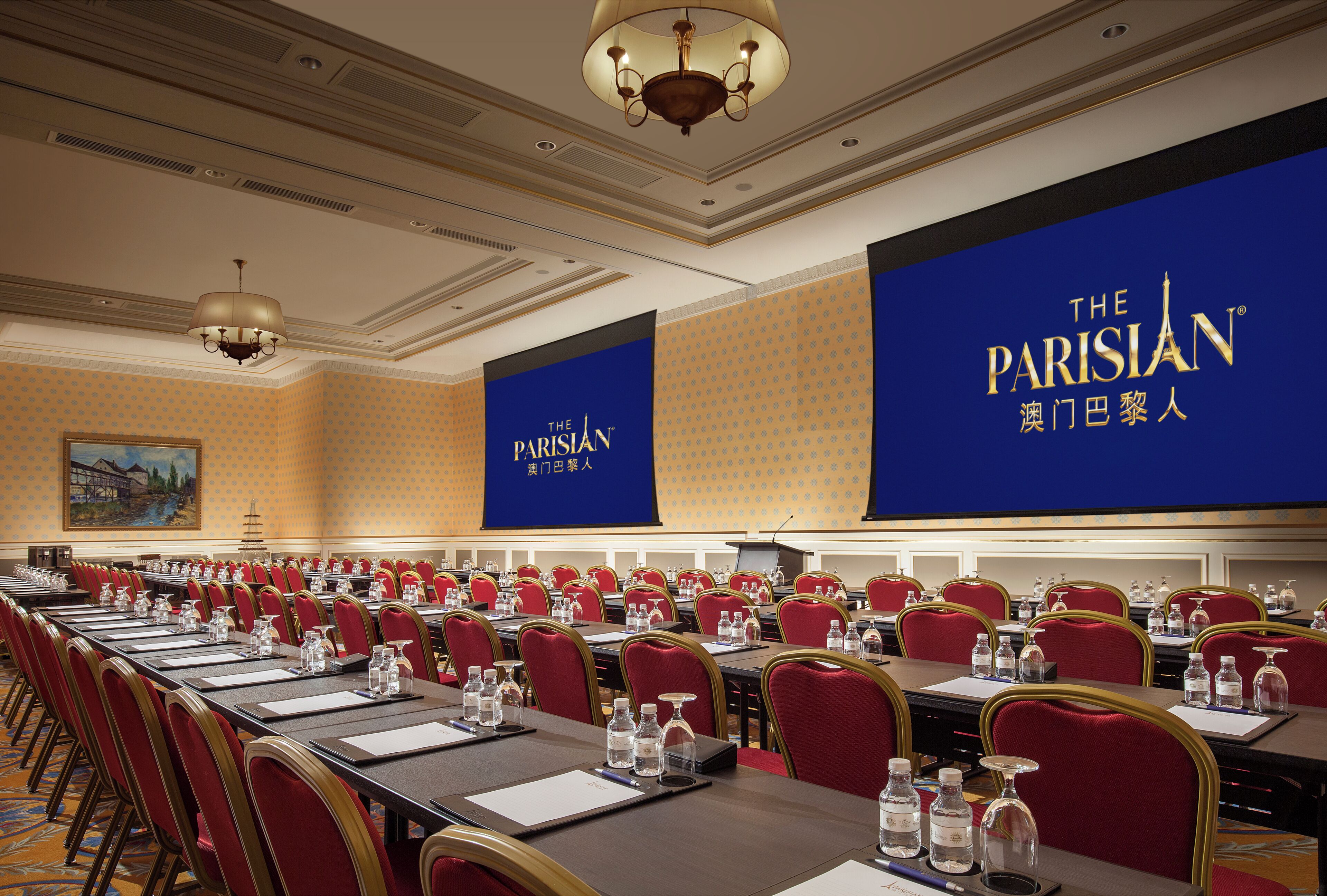 A conference room with rows of red chairs, tables with water bottles, and a large screen displaying 'The Parisian' branding.