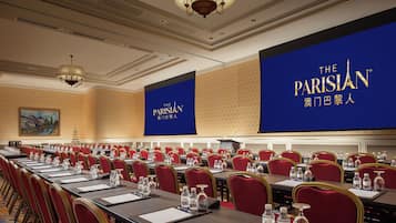 A conference room with rows of red chairs, tables with water bottles, and a large screen displaying 'The Parisian' branding.