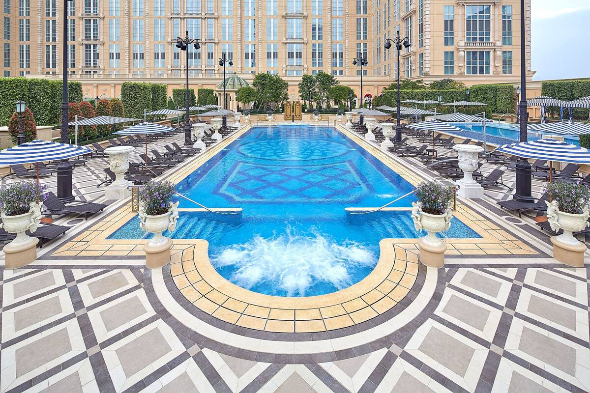 A large indoor swimming pool with a geometric tile design, surrounded by lounge chairs and umbrellas, with a large building in the background.