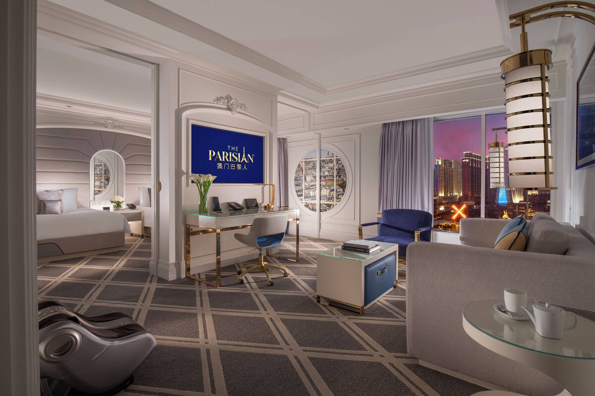 A modern hotel room with a city view, a desk with a computer, a massage chair, a sofa, and a glass coffee table.