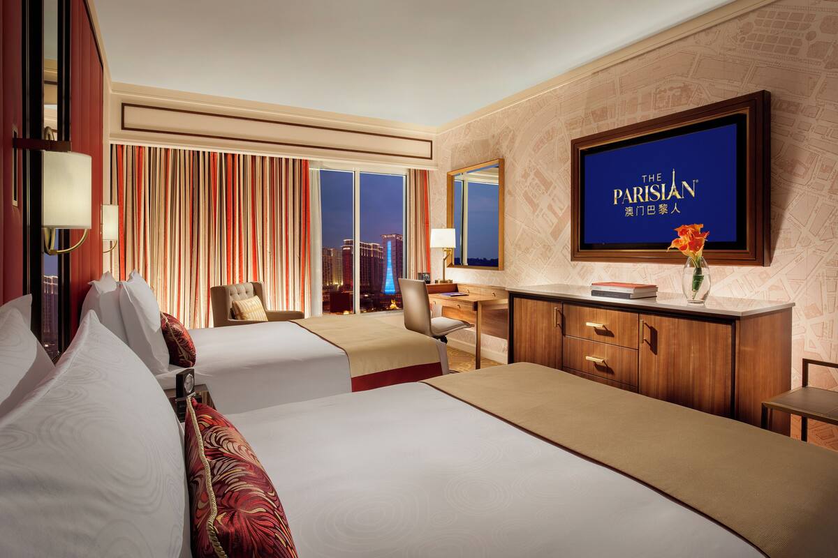 A hotel room with two beds, a desk, a chair, a TV, and a view of the cityscape.