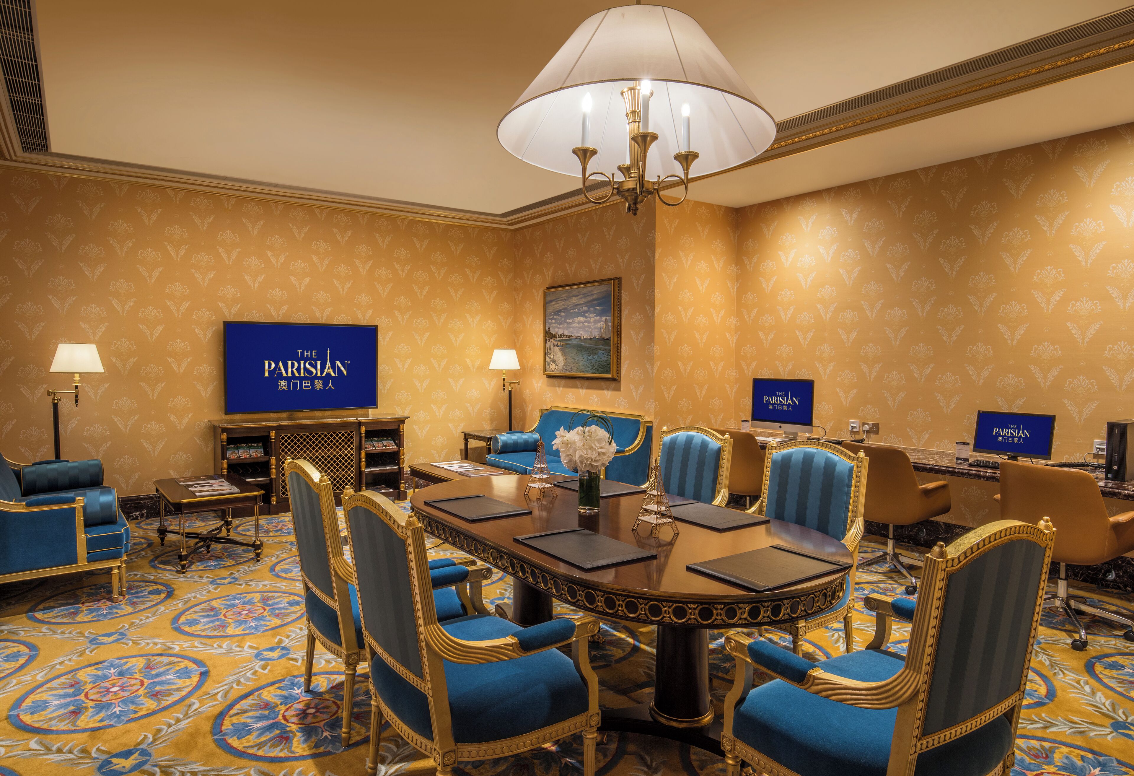 Een vergaderruimte met een grote tafel, blauwe gestoffeerde stoelen, een kroonluchter en een bord met de tekst 'The Parisian' aan de muur.