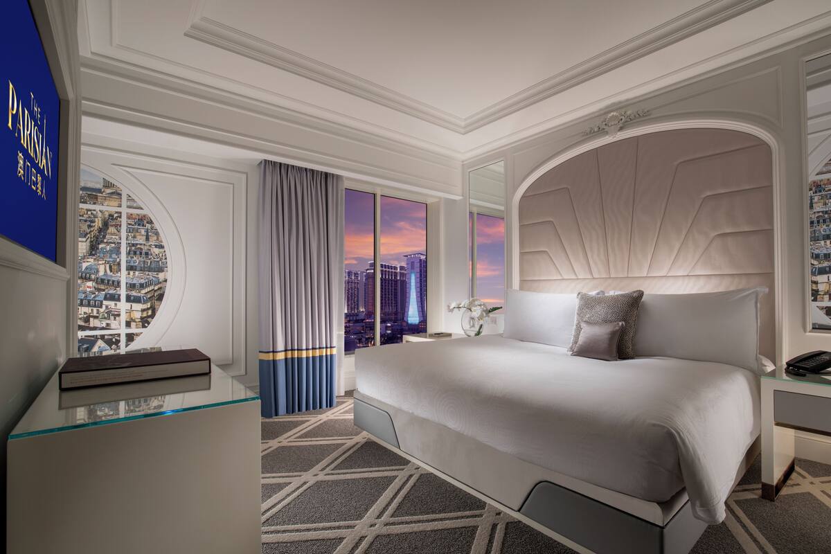 A modern hotel room with a large bed, a glass desk, a city view, and a decorative headboard.