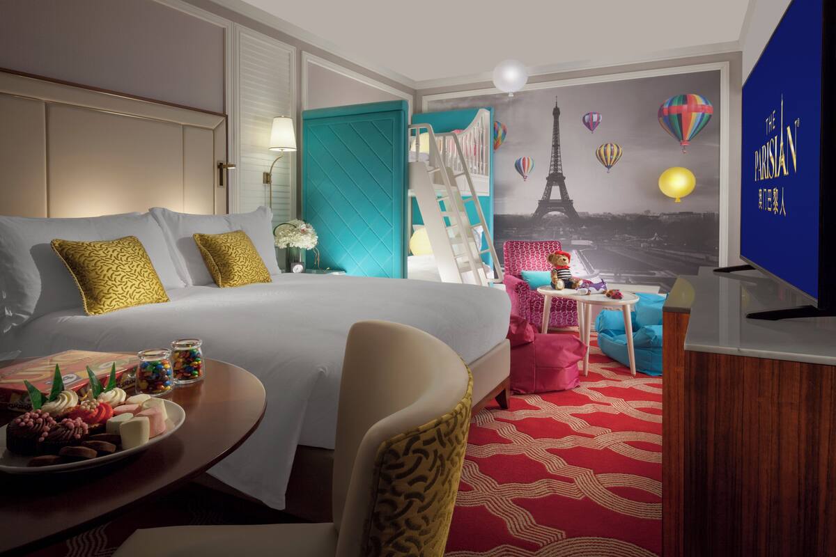 A hotel room with a bed, a desk with a chair, a TV, and a wall mural of Paris.