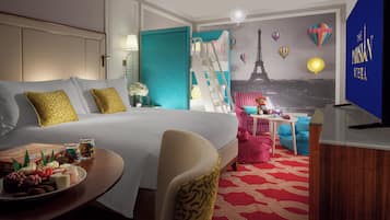 A hotel room with a bed, a desk with a chair, a TV, and a wall mural of Paris.