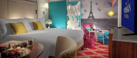 A hotel room with a bed, a desk with a chair, a TV, and a wall mural of Paris.