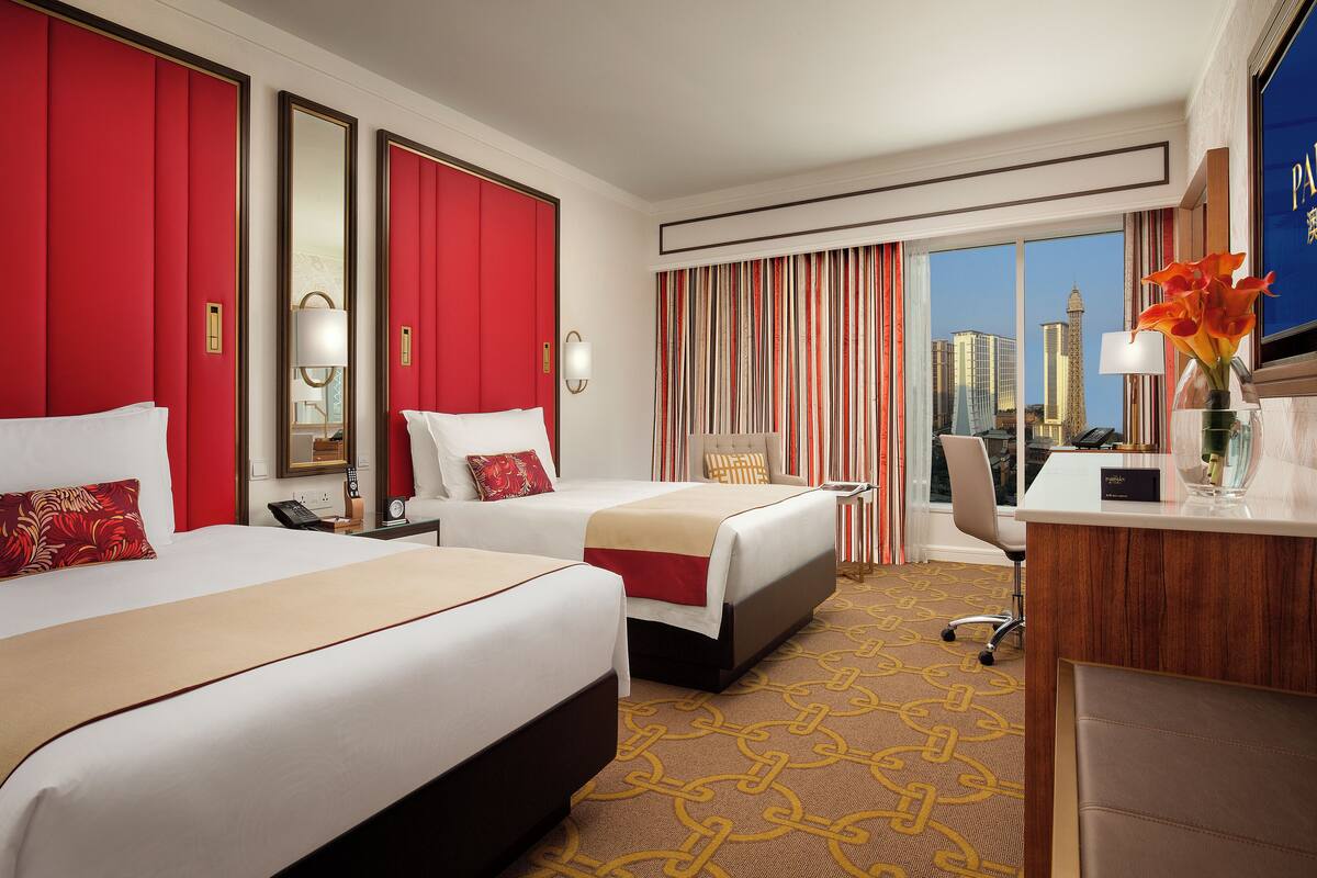A hotel room with two beds, a desk, a chair, a phone, and a view of the cityscape.