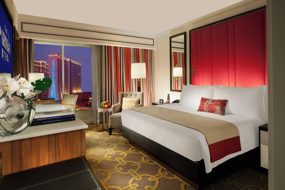 A hotel room with a bed, a desk with a laptop, a chair, a nightstand, and a large window with a city view.