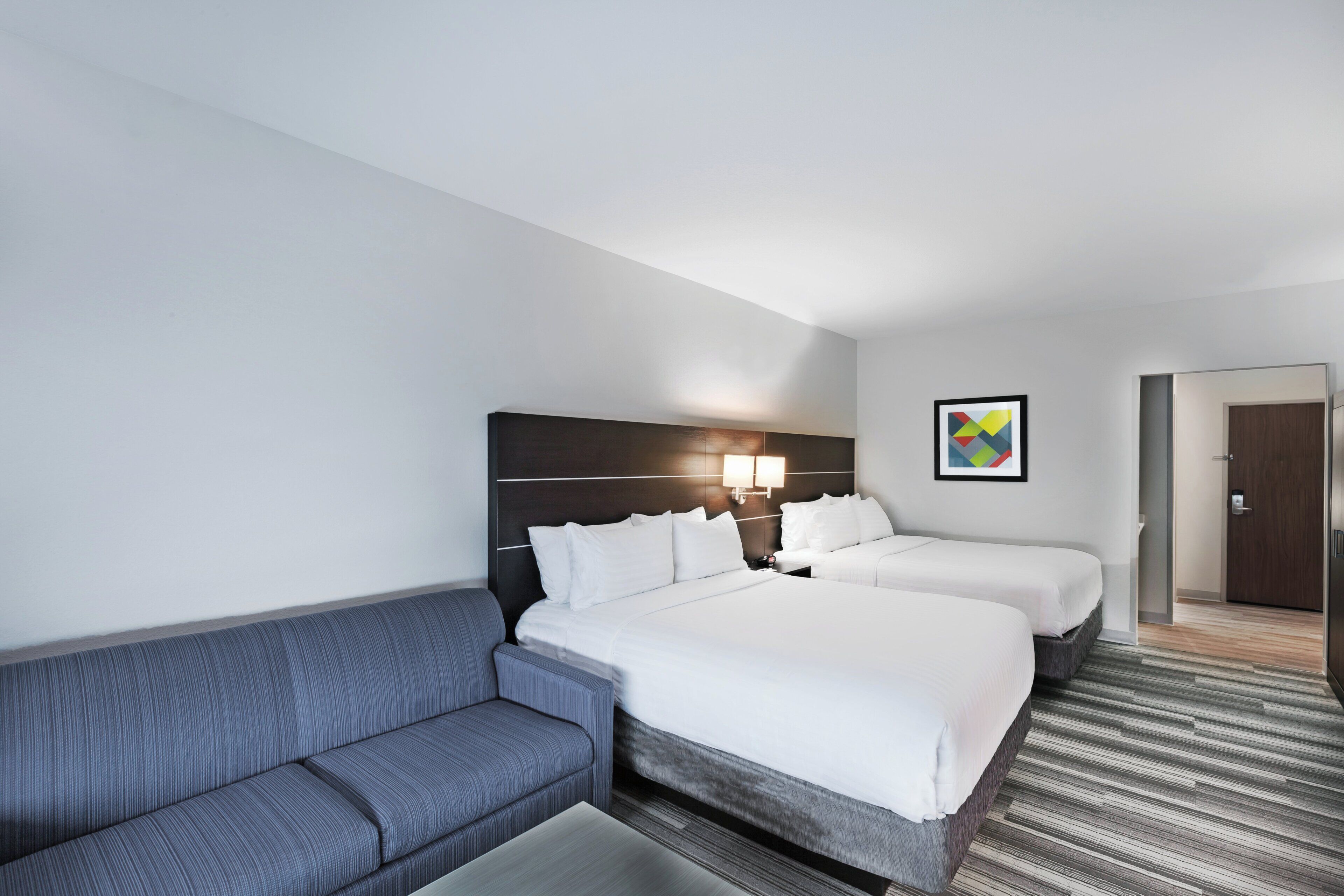 Habitación de hotel con dos camas, un sofá, una lámpara de pared y una puerta.