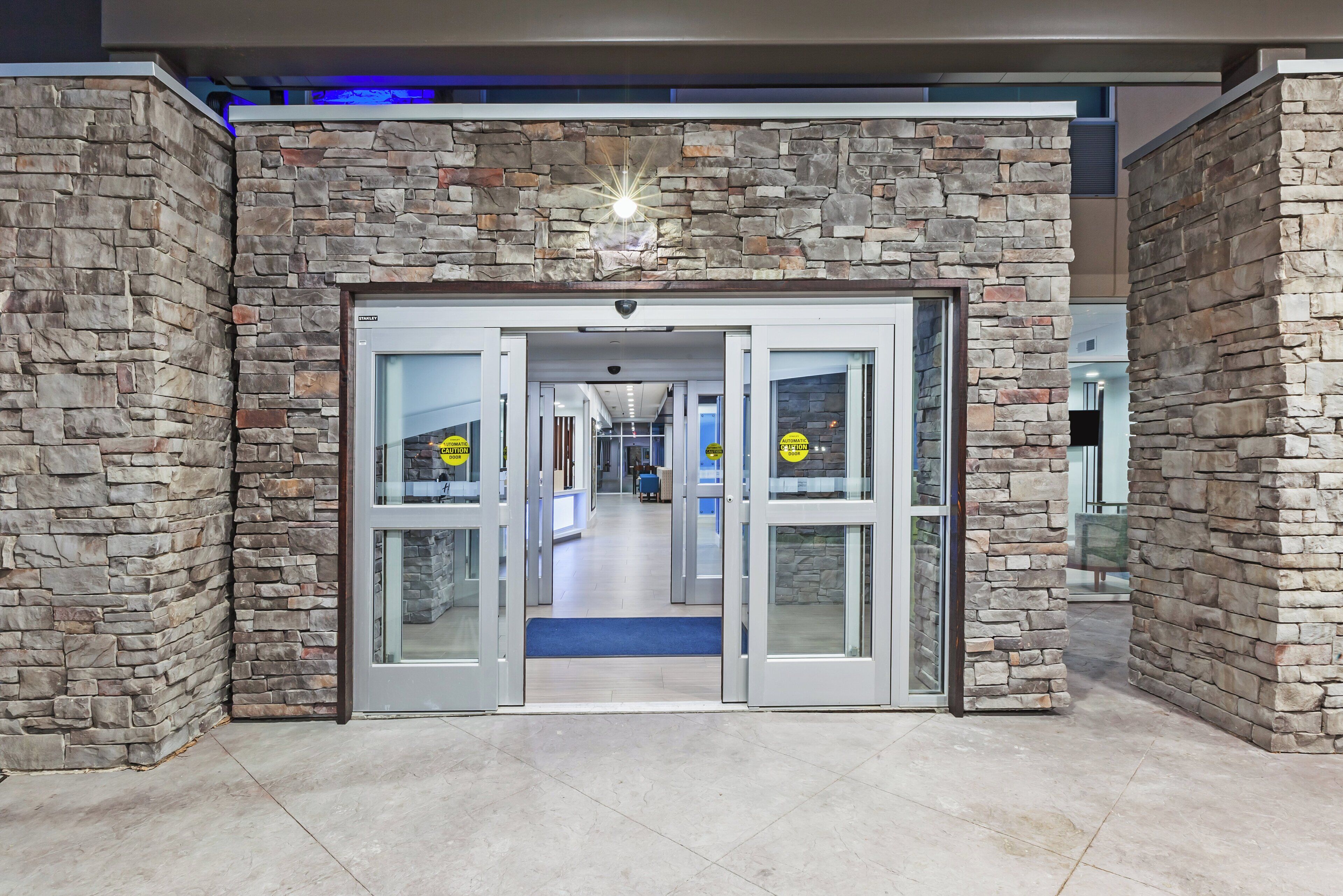 Entrada de un hotel con fachada de piedra, puertas automáticas y una alfombra azul que conduce al interior.