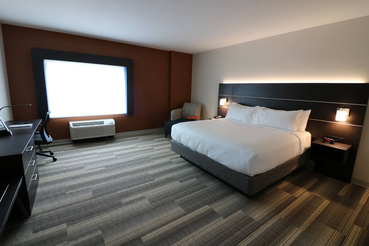 A modern hotel room with a large bed, a desk, and a chair. There is a window providing natural light and a wall-mounted air conditioner.