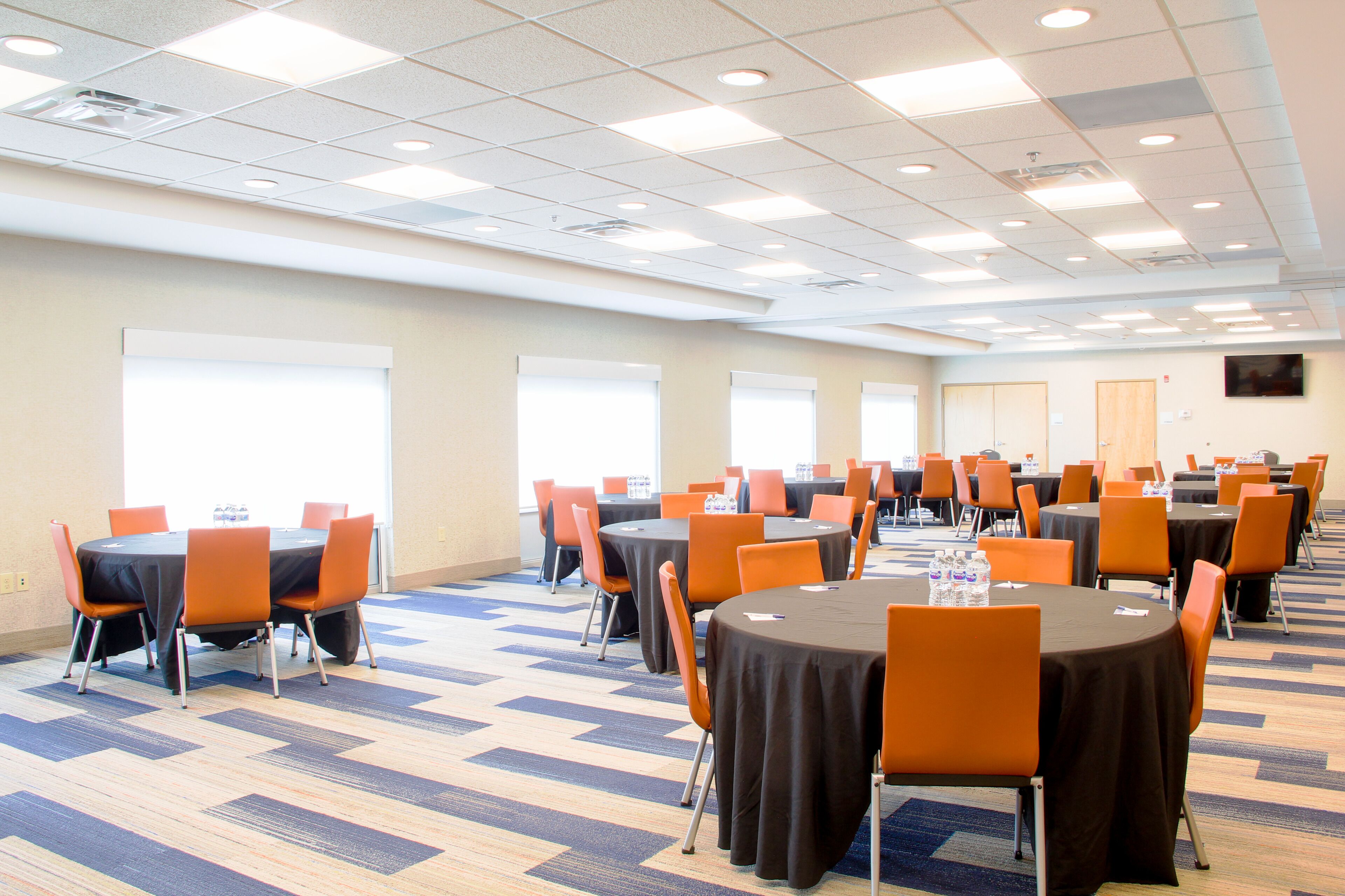 A conference room with multiple tables and chairs, arranged in rows, with a carpeted floor and a ceiling with recessed lighting.
