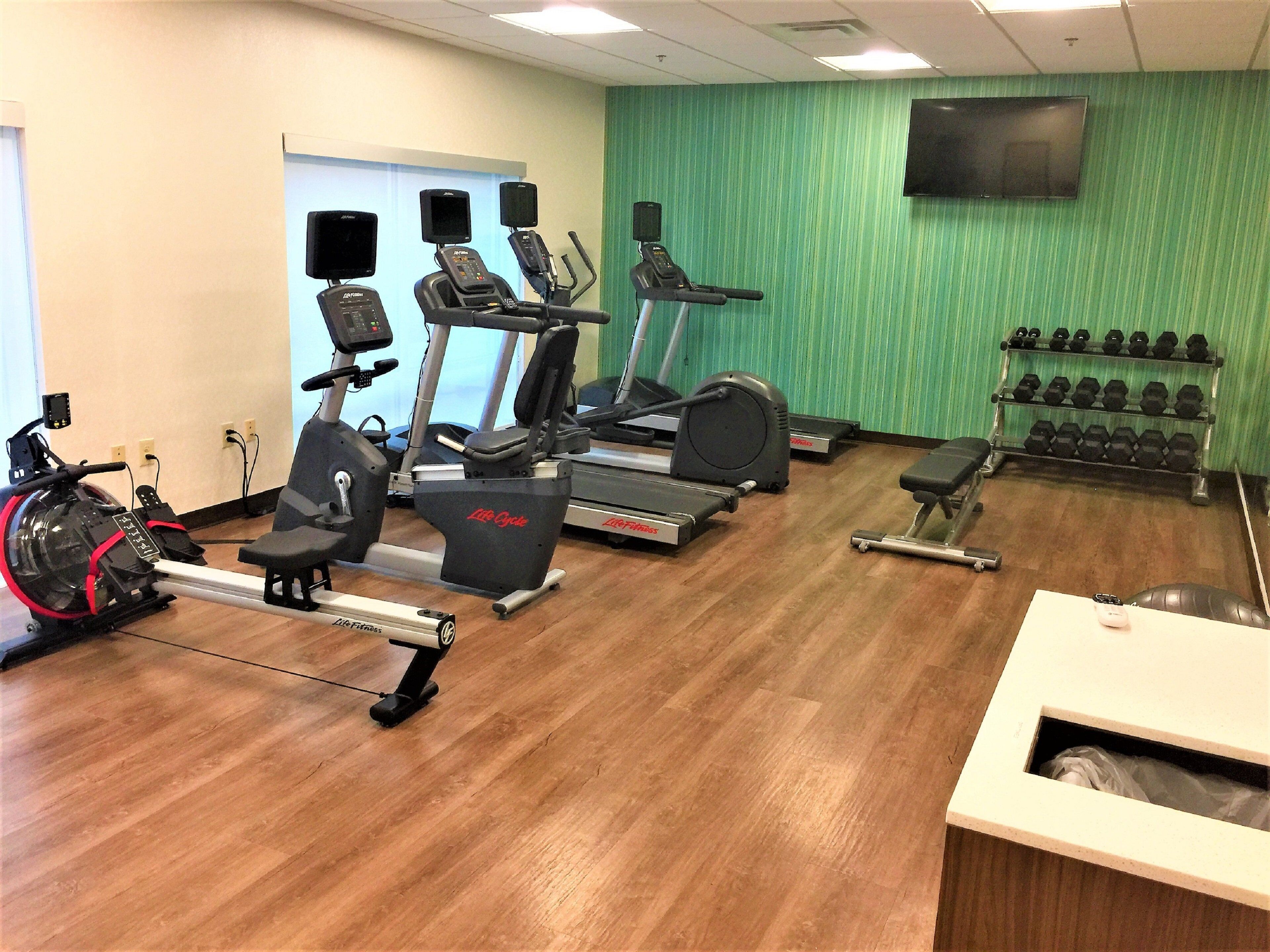 A gym with cardio machines, dumbbells, and a TV on the wall.
