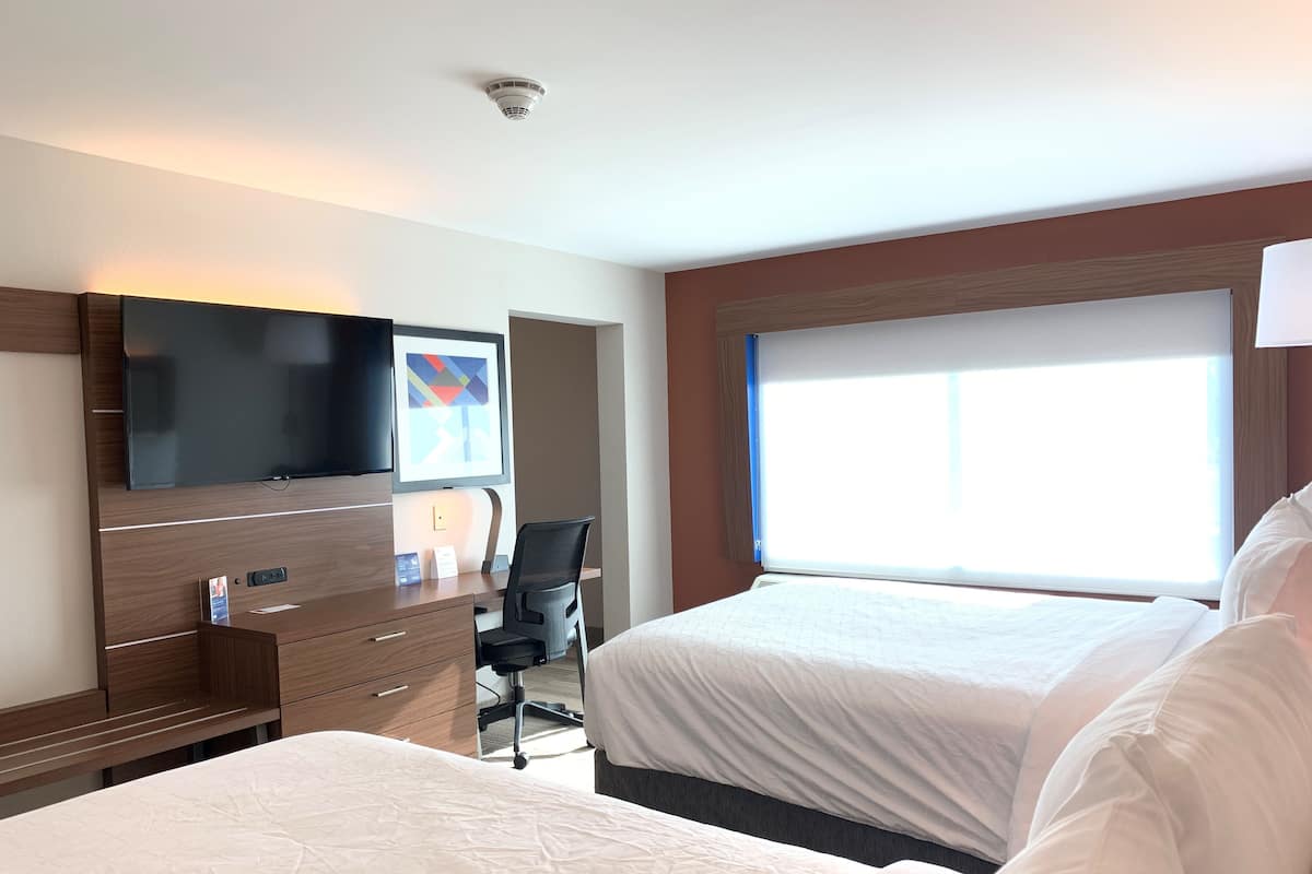 A hotel room with two beds, a desk with a chair, a flat-screen TV, and a large window.