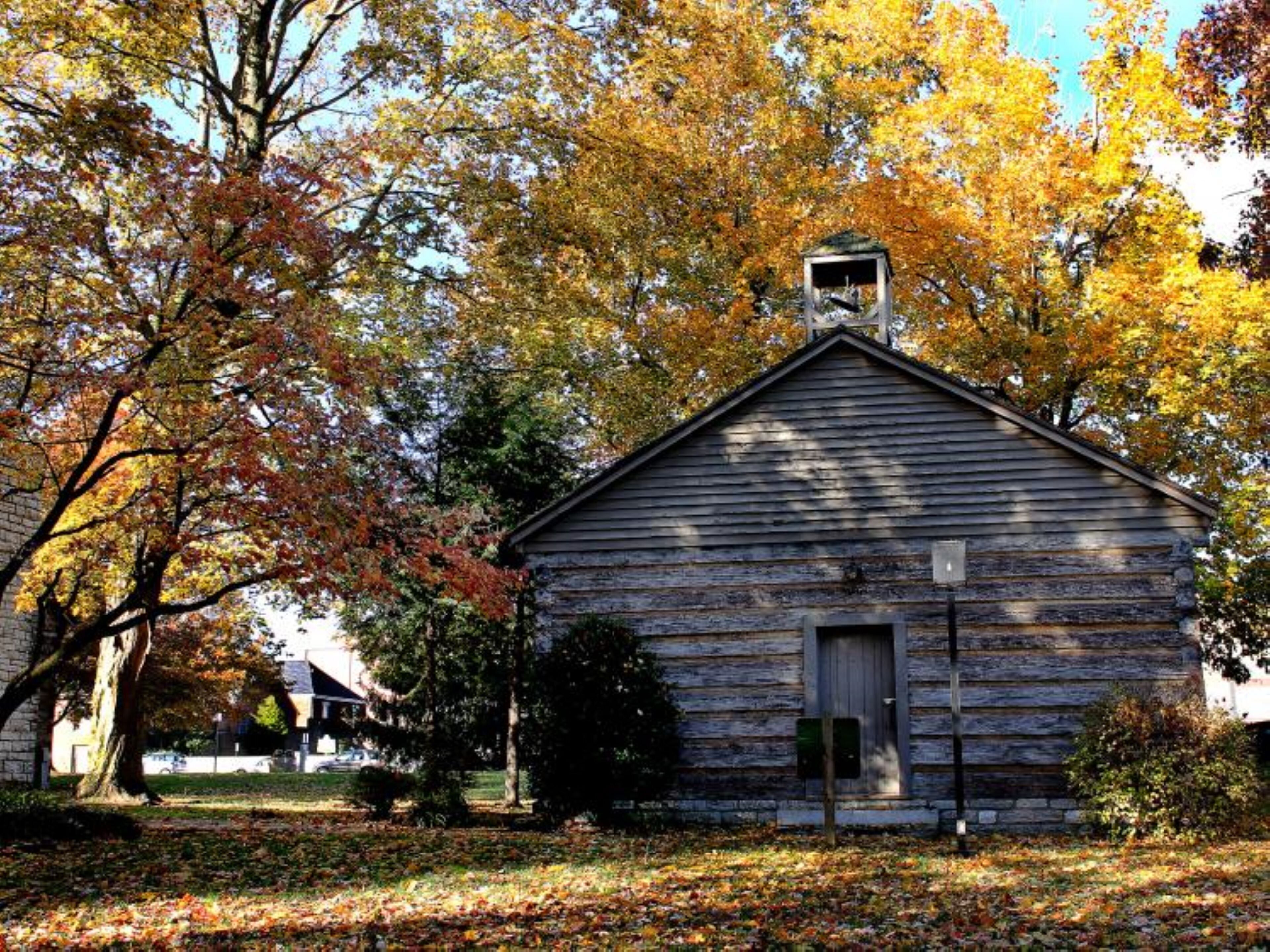A rustic wooden cabin with a gabled roof, surrounded by trees with autumn foliage.
