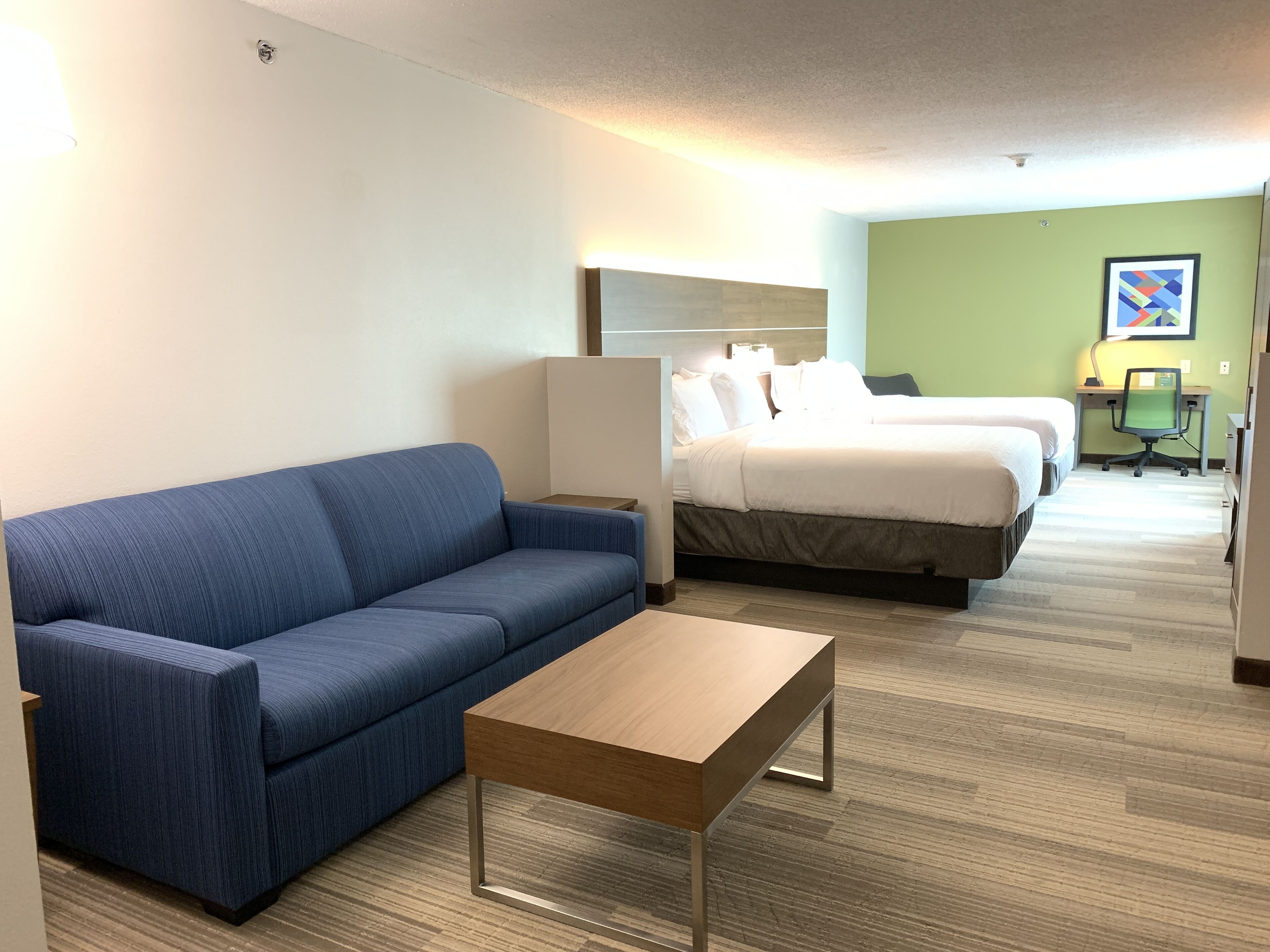 A hotel room with a blue sofa, a wooden coffee table, two beds, a desk with a chair, and a painting on the wall.