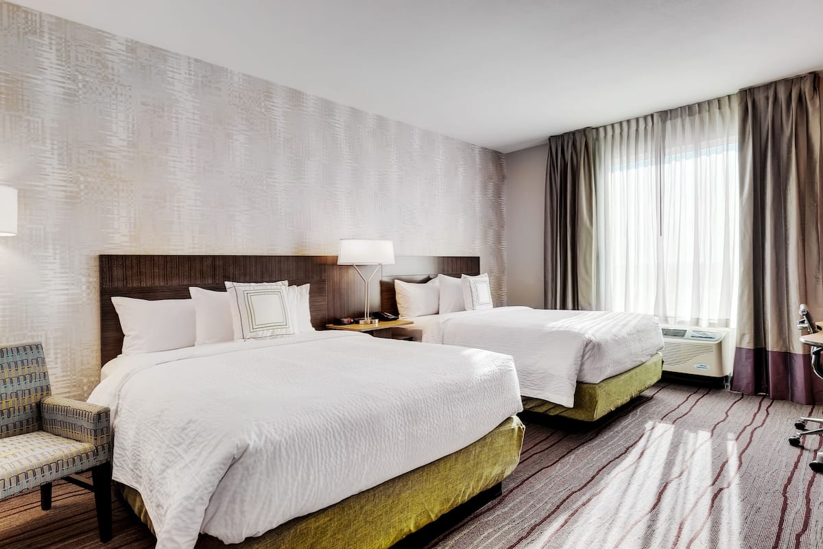 A hotel room with two beds, a nightstand, a lamp, a chair, and a large window with curtains.