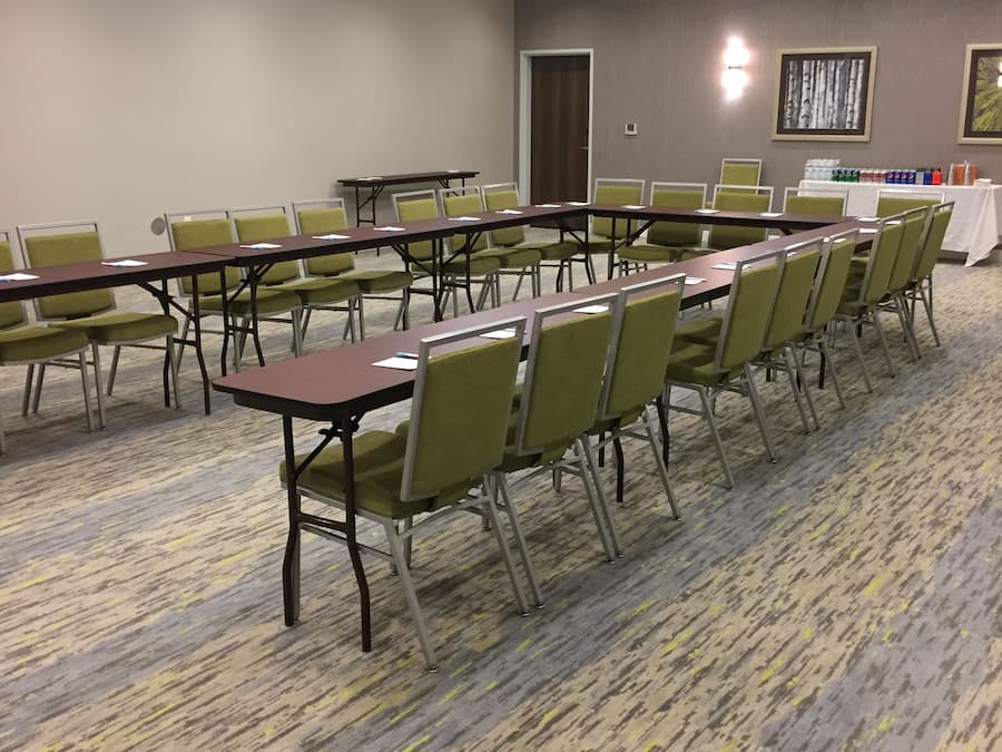A conference room with long tables and green chairs, carpeted floor, and ceiling lights.