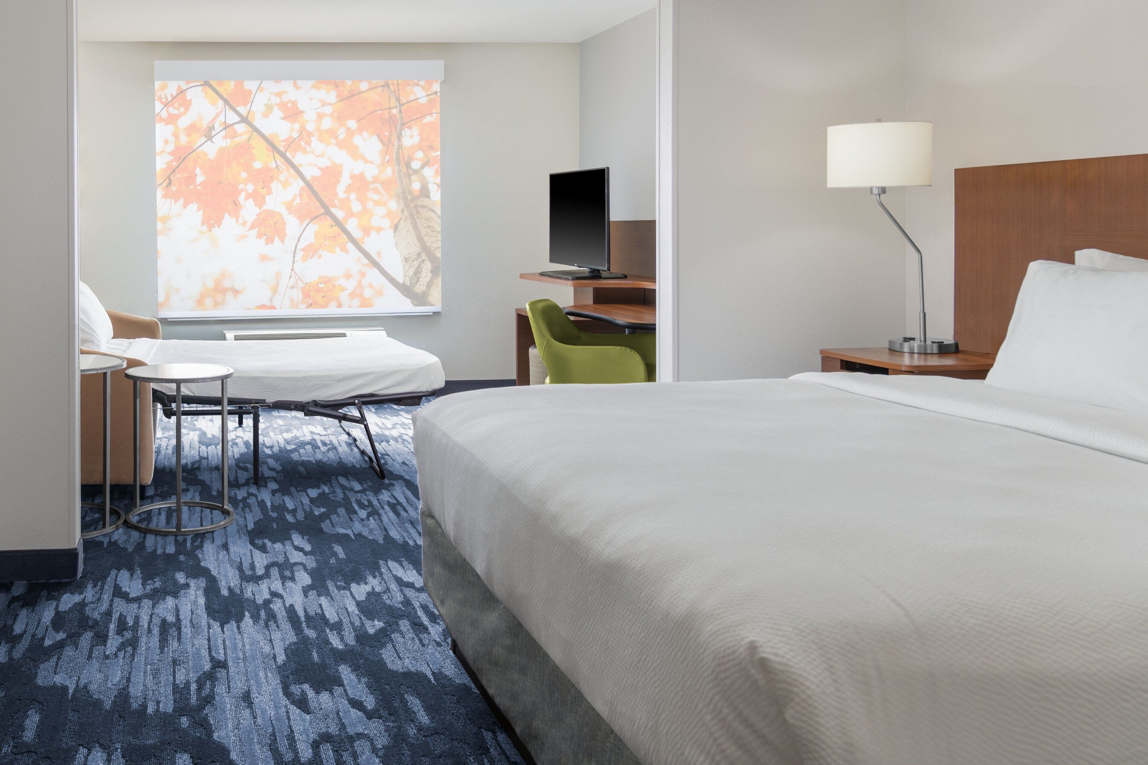 A hotel room with a large bed, a desk with a computer, a chair, a lamp, and a window with a view of trees.