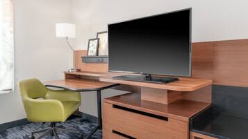 A hotel room with a desk, a green chair, a flat-screen TV, a lamp, and framed pictures on the wall.