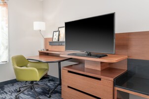 A hotel room with a desk, a green chair, a flat-screen TV, a lamp, and framed pictures on the wall.