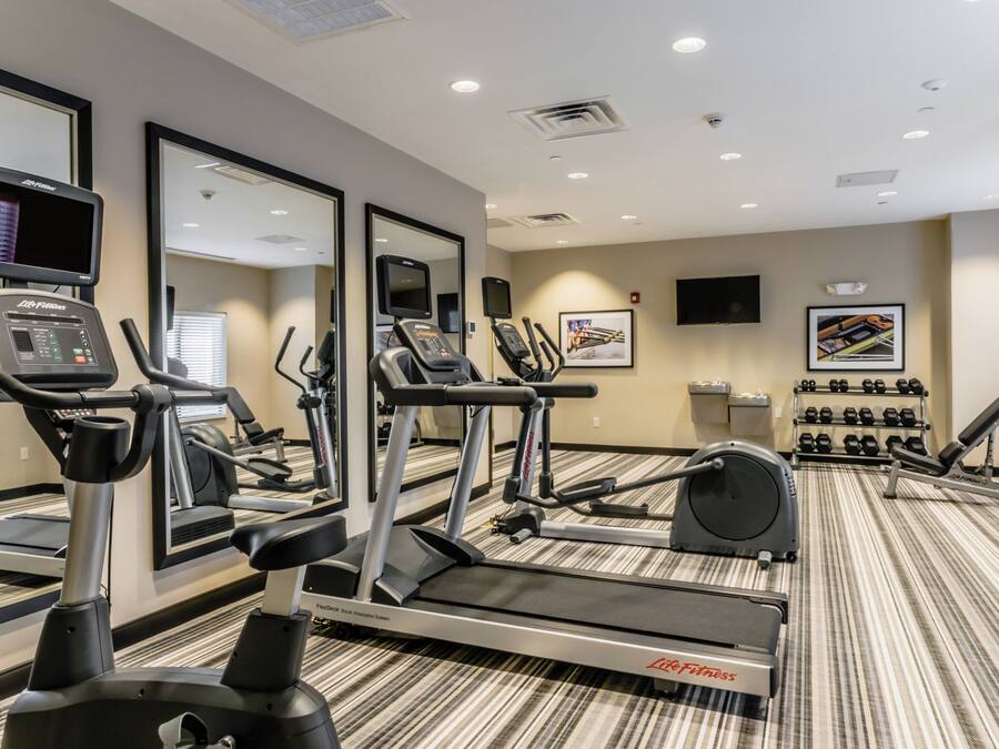 Un gimnasio con diversas máquinas de cardio, espejos, un televisor y mancuernas.