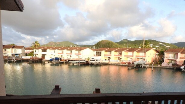 A marina with docked boats and a row of houses with balconies, overlooking a calm body of water and hills in the background. - Villa 230C at Jolly Harbour (Jolly Harbour)