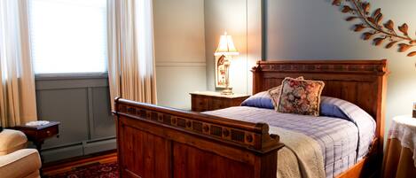 Una cama de madera con cabecera, una mesita de noche con lámpara, una silla, una ventana con cortinas y un adorno de pared.