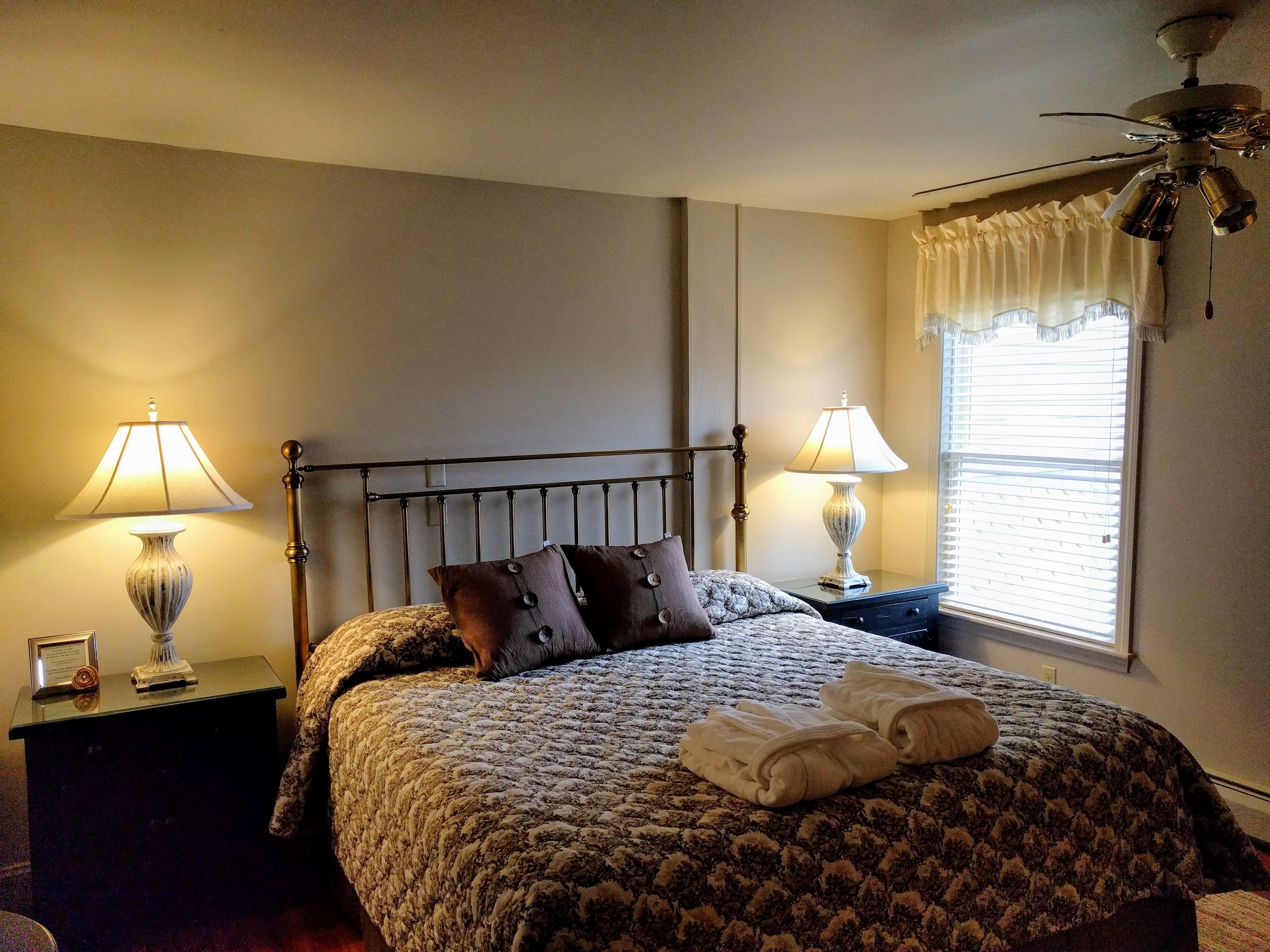 A neatly made bed with a patterned bedspread, two bedside lamps, a ceiling fan, and a window with blinds.