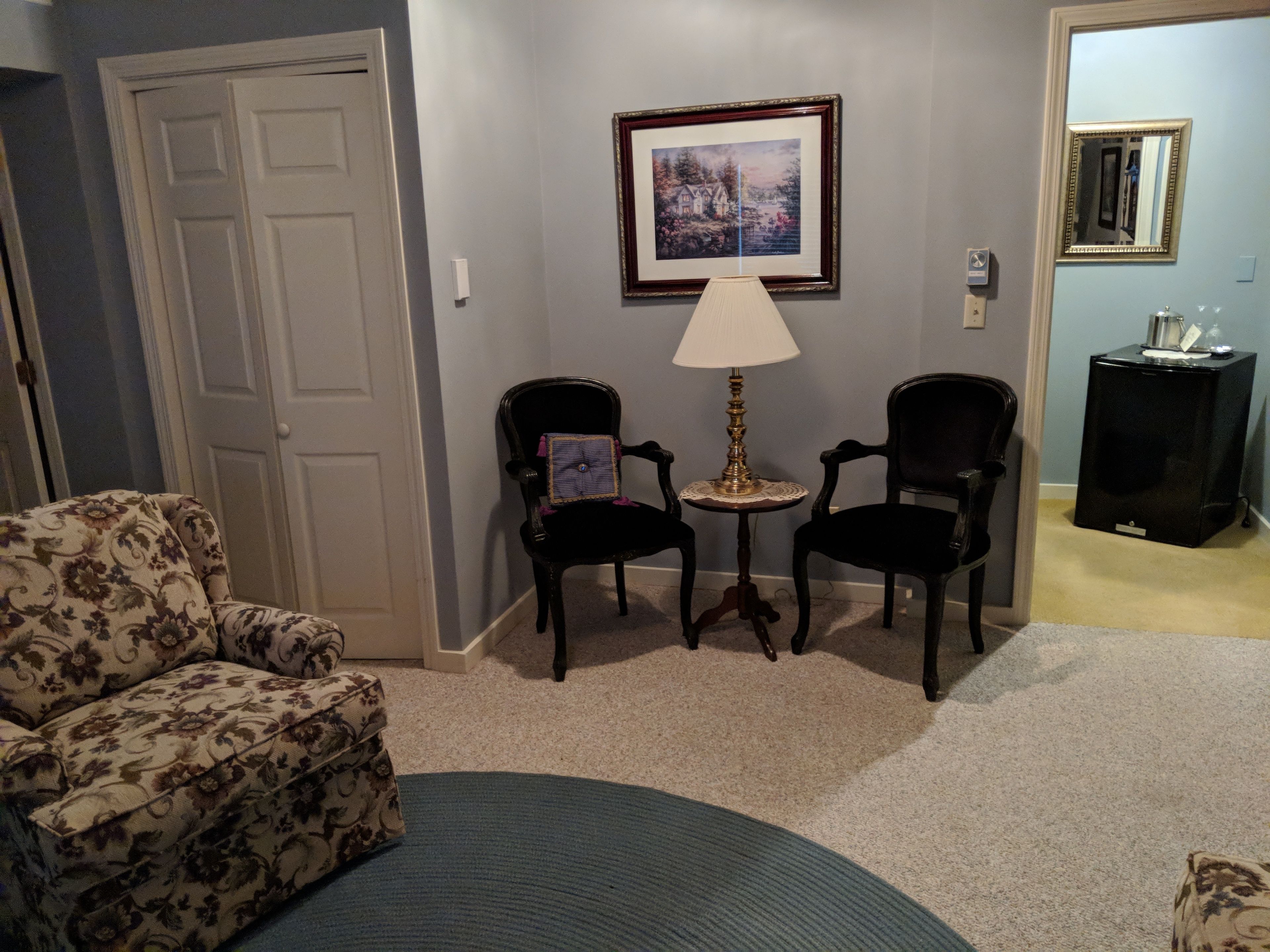 A living room with a floral-patterned sofa, two dark chairs, a small round table with a lamp, a framed picture on the wall, and a black side table with a decorative item.