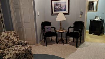 A living room with a floral-patterned sofa, two dark chairs, a small round table with a lamp, a framed picture on the wall, and a black side table with a decorative item.