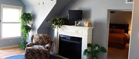 A room with a fireplace, a ceiling fan, a TV mounted above it, a bed with a headboard, and a large window.