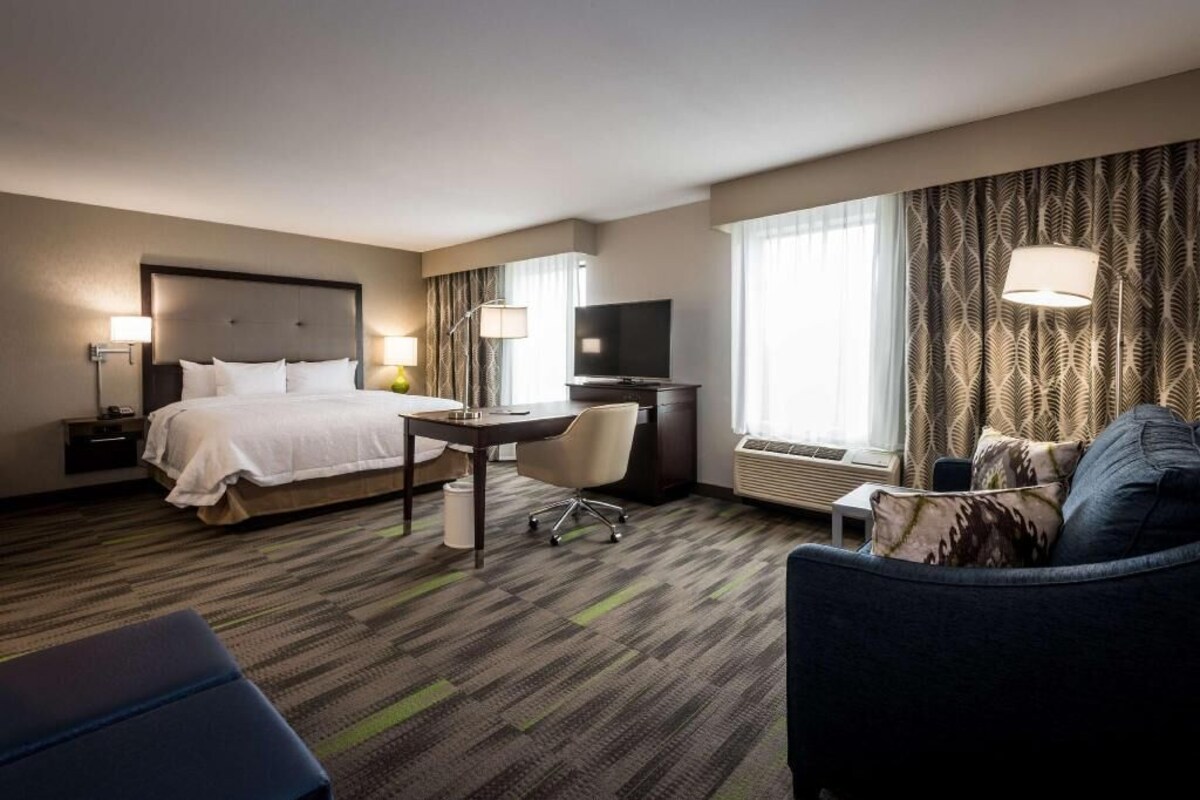 A hotel room with a bed, desk, chair, TV, and large windows.