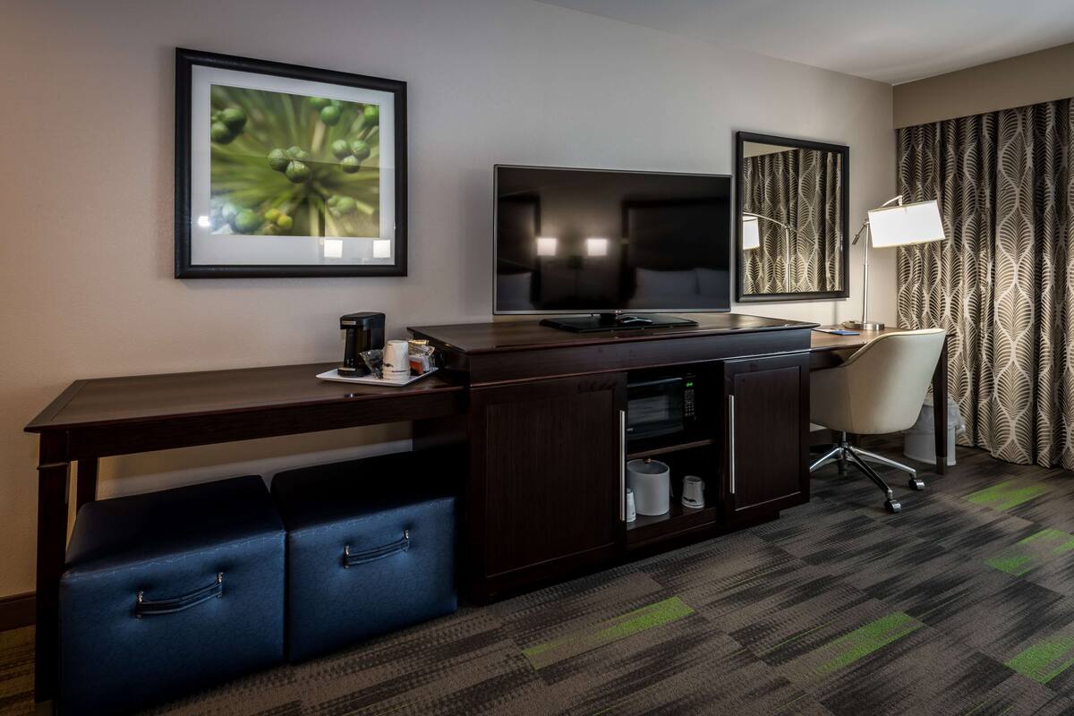 A hotel room with a dark wood desk, a flat-screen TV, a large mirror, a desk lamp, a chair, and a patterned carpet.