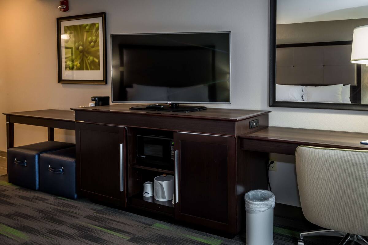 A hotel room with a dark wood desk, a flat-screen TV, a microwave, a mini-fridge, a trash bin, and a large mirror.