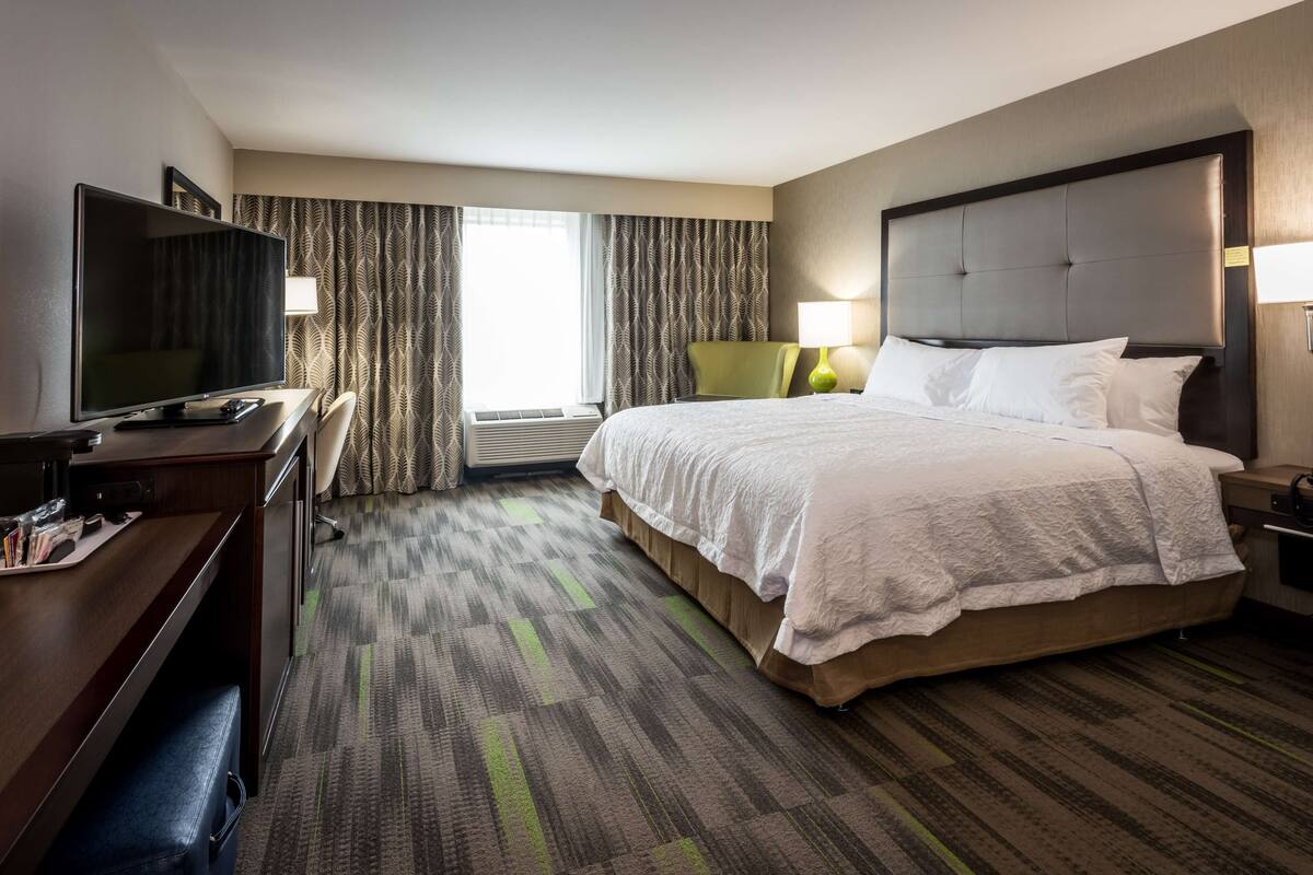 A hotel room with a bed, a desk with a TV, a chair, and a window with curtains.