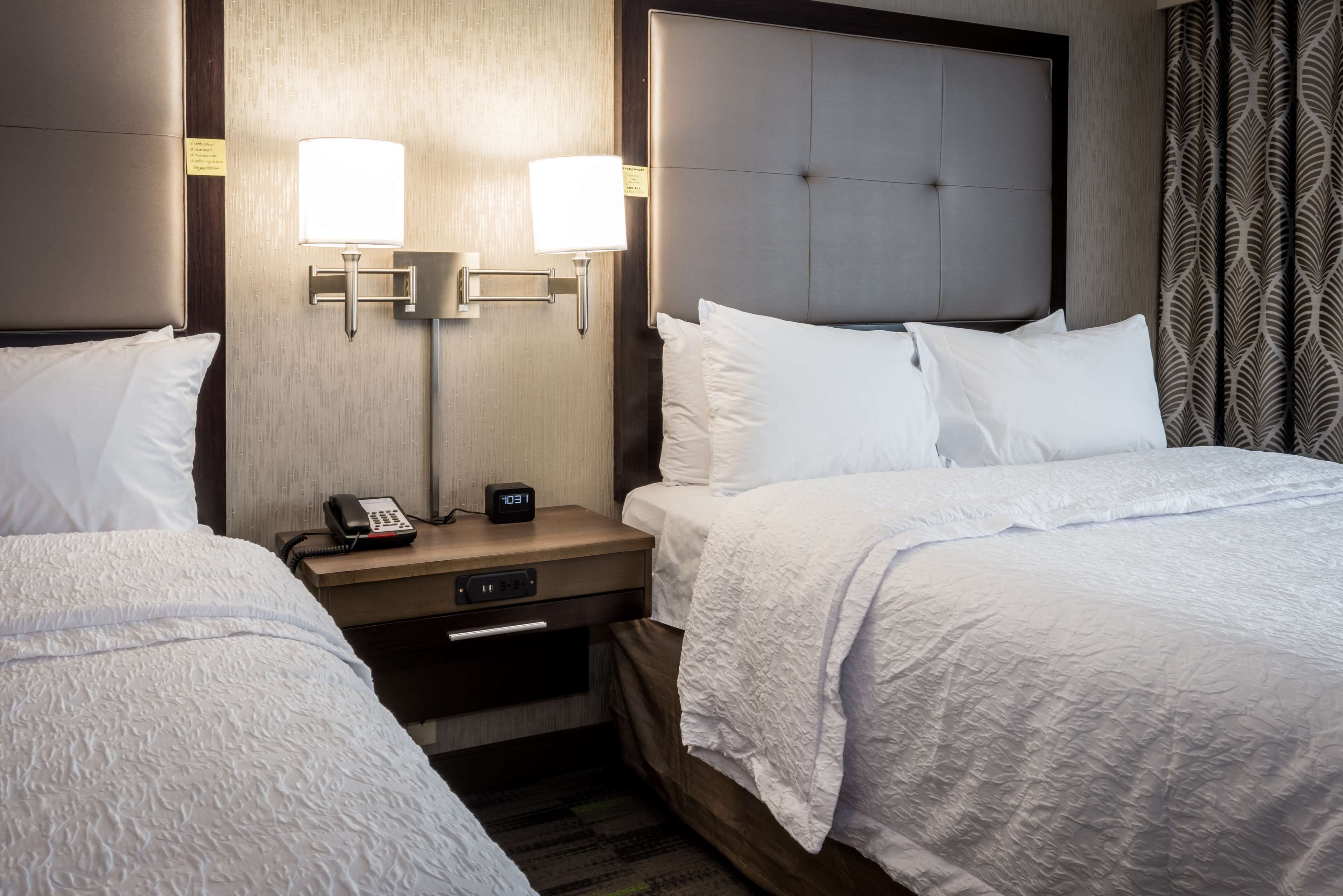 A hotel room with two beds, a nightstand with a phone and clock, and wall-mounted lamps.