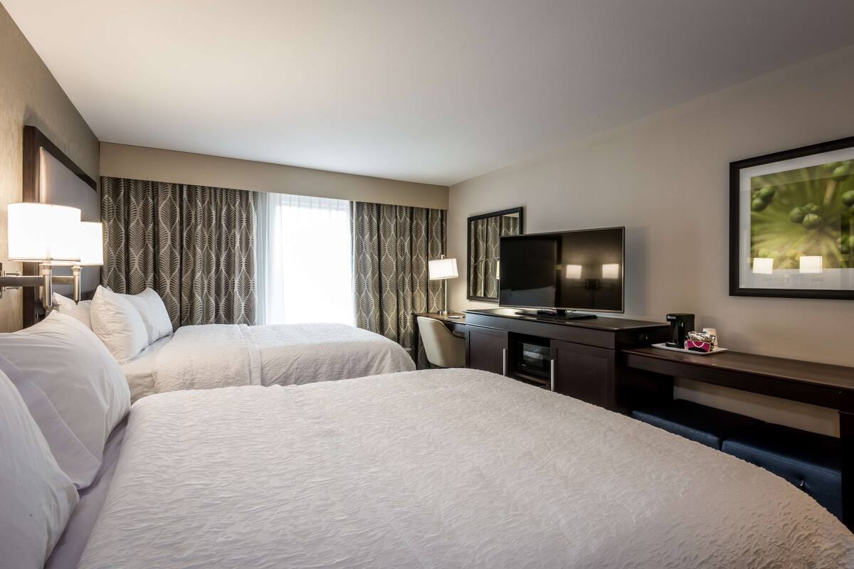 A hotel room with two beds, a desk with a television, and a large window with curtains.